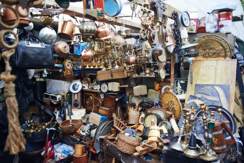 A dense display of various used household items and decorative objects, including copper and brass fittings, small furniture pieces, clocks, and picture frames, all arranged on tables and shelves. The collection features a mix of textures and finishes, with shiny metallic surfaces, aged wood, and fabric textiles visible among the clutter. Items are stacked and grouped together, with some hanging or leaning against each other, creating a busy and cluttered scene typical of second-hand markets or clearance yard sales. The environment appears to be outdoors or in an open-fronted setting, with natural daylight illuminating the assortment. This scene relates to rubbish removal services, such as those provided by Office Clearance Leytonstone, which often deal with clearing similar collections of unwanted possessions from residential or commercial premises in Leytonstone or nearby areas. The overall impression is of a large variety of discarded or pre-owned objects awaiting disposal or resale within a thorough clearance process.