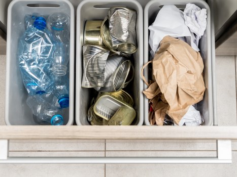 The image shows a close-up of three grey plastic recycling bins positioned on a light tiled floor, likely outside a commercial or domestic setting. The left bin contains several clear plastic bottles, some with blue caps, with the bottles being empty and crushed. The middle bin holds a cluster of opened and flattened aluminium cans, with some stacked and overlapping, their metallic surfaces reflecting light and showing ridges and embossed patterns typical of beverage cans. The right bin contains crumpled paper and a large piece of brown kraft paper, both appearing used and loosely folded. The bins are placed directly on the tiled ground, which is clean and uniform in color. This scene is representative of waste collection or rubbish disposal practices, possibly related to household or small business rubbish management, relevant to Leytonstone's local waste removal services, especially in an urban or suburban environment. The overall lighting suggests natural daylight, with no other background elements visible.