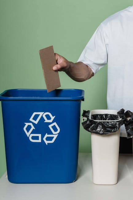 A person’s hand holding a semi-transparent light blue plastic trash bag, which appears to be filled with waste materials. The bag is gathered at the top and tied shut, with the creases and folds of