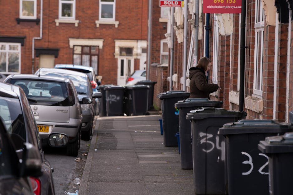 A black plastic wheeled rubbish bin labeled 'St. John's' positioned on a sidewalk at night, with its lid open and filled with various waste items, including cardboard boxes, paper, and black plastic b