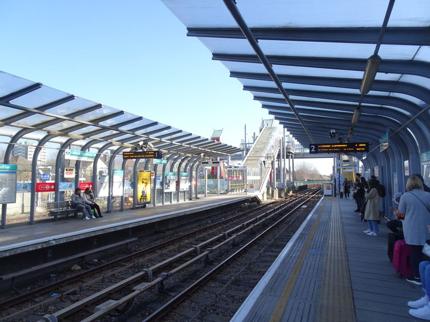 A modern, covered railway station platform in daylight with a curved, transparent roof supported by metal beams. On the left side, a waiting area with two people sitting on a bench beneath the timetab