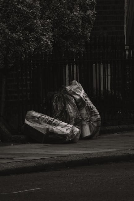 Two large black plastic rubbish bags filled with waste material are placed on the pavement beside a wooden fence and a tree, with a brick wall and foliage in the background. The bags appear to be tigh