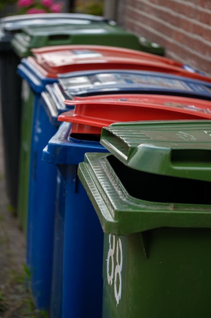 A person's arm is extended into the frame holding a green plastic bag filled with waste material. The bag appears slightly transparent, revealing light-colored and darker green trash objects inside. T