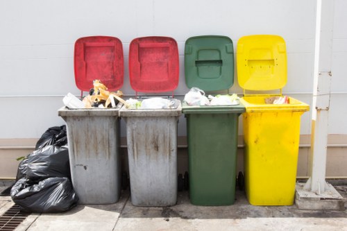 The image displays four large wheelie bins positioned side by side outdoors on a concrete pavement against a light grey wall. From left to right, the bins are colored red, red, green, and yellow, with their lids open, revealing mixed waste including plastic packaging, cardboard, and other household rubbish. To the left of the grey bins, there are several black garbage bags stacked, placed directly on the pavement. The surface of the bins shows signs of wear and dirt, typical of outdoor rubbish collection points. The environment appears clean and orderly, with no visible signage or markings. The arrangement suggests a designated waste disposal area, possibly part of an office or commercial premises, consistent with services offered by a company like Office Clearance Leytonstone for rubbish removal and waste management. The scene captures a typical outdoor rubbish collection setup, emphasizing the importance of appropriate waste segregation and disposal within local urban environments such as Leytonstone and surrounding areas.
