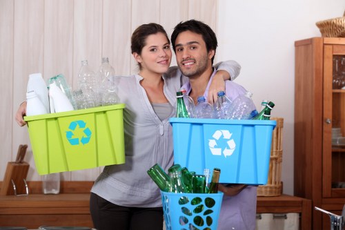 A young couple standing indoors in a room with light wooden paneling on the walls, each holding a plastic recycling bin filled with glass bottles; the woman has long dark hair and is wearing a light gray top, while the man has dark hair and facial hair, dressed in a light purple shirt. The green bin held by the woman contains clear glass bottles, while the blue bin held by the man is filled with various clear and green glass bottles. A smaller blue basket in front of them holds additional green glass bottles. In the background, there is a wooden sideboard with some decorative items and a woven basket, and the overall lighting suggests a well-lit interior environment. This image visually relates to waste disposal and recycling services that might be offered by Office Clearance Leytonstone, aligned with domestic glass waste collection in the Leytonstone area.