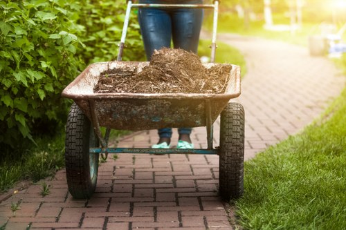 A person stands behind a two-wheeled wheelbarrow filled with dark, moist soil or garden waste, positioned on a paved sidewalk adjacent to a grassy area. The person’s legs are visible, dressed in blue jeans, with the upper body and face not shown. The wheelbarrow has black rubber tires and a metal frame, with a rust-colored, weathered basin containing the soil. To the left, there is a dense hedge of green foliage, and in the background, part of a residential street or garden path is visible, illuminated by sunlight. The setting appears to be outdoors in a suburban or residential environment, suitable for waste removal or garden clearance services in Leytonstone, as handled by Office Clearance Leytonstone.