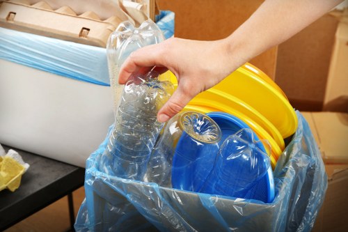 A close-up of a person's hand holding a clear plastic bottle filled with transparent liquid, positioned above a large black bin liner containing various coloured plastic containers, including yellow and blue buckets and lids, in an indoor setting. The bin liner is partly open and placed on a dark surface, with a white plastic storage container and cardboard boxes visible in the background. The scene suggests the collection of recyclable plastic waste, indicative of rubbish removal and waste sorting activities typical of office clearance services in Leytonstone. The environment appears well-lit, highlighting the textures of the plastic objects and the smooth surface of the bottle.